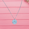 A sterling silver necklace with a circular pendant featuring an ocean wave design, displayed against a pink wooden background.